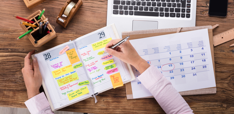 Close-up Of Businesswoman Writing Schedule In Calendar Diary On Desk