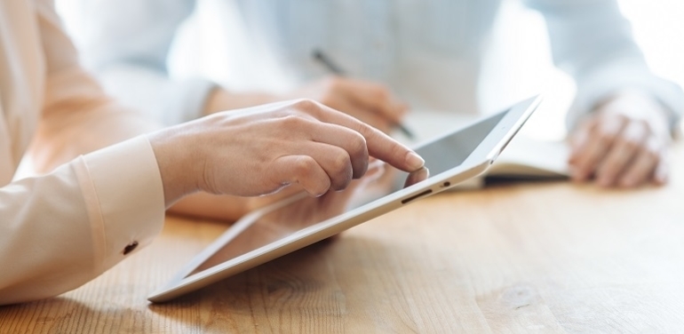 Two female businesswomen working their hands on a tablet in an office in the sunlight at a wooden table and one of them writes a pen in a stylish notebook, and the other touches the tablet with her finger