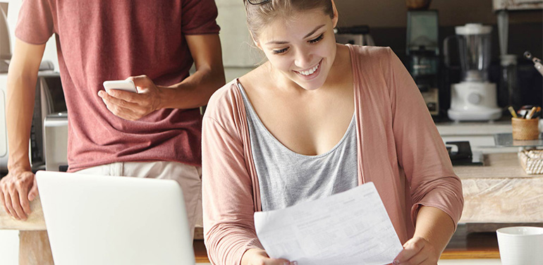 young woman sitting at a table, smiling looking at a piece of paper, her partner is standing in the background