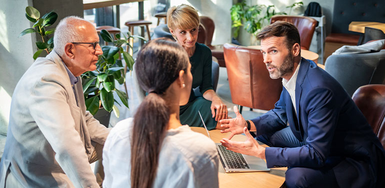 two men and two women sit around a table, looking at one man discussing business