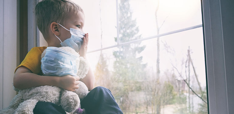 young child holding a teddy bear, both wearing face masks looking out the window