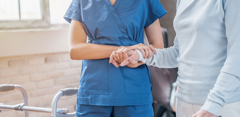 female nurse supporting patient to walk, photo shows two people from the neck down, nurse has her hand on patient's arm