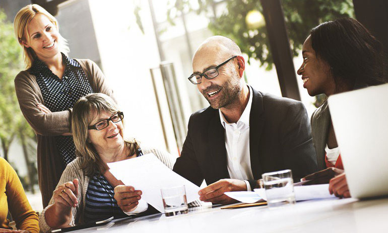 group of colleagues at a table smiling, a man in the center of the picture is reading a piece of paper