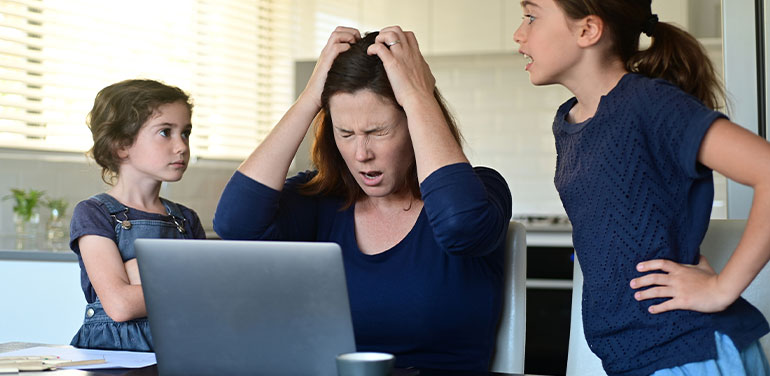 A woman trying to work on her laptop, holding her head in her hands, two young girls arguing next to her