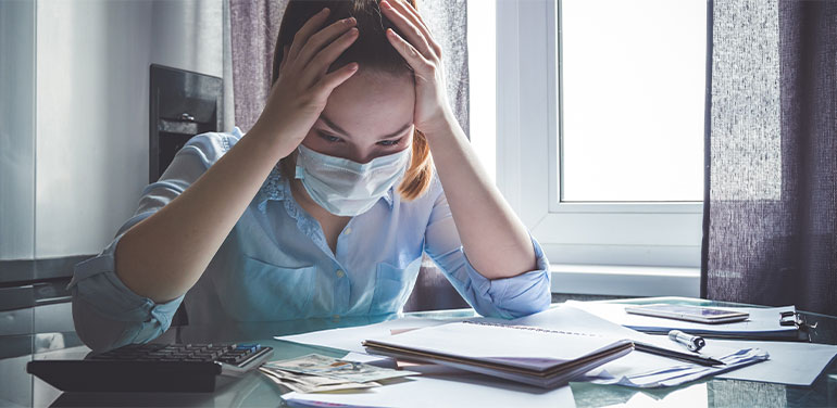 Woman sat at a table wearing a facemask with her head in her hands looking at paperwork and a calculator