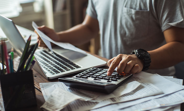 close up of man working out something on a calculator while holding a piece of paper in his other hand, with a laptop in front of him on the table