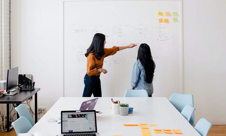 two women pointing at a whiteboard, with a table and a laptop in the foreground