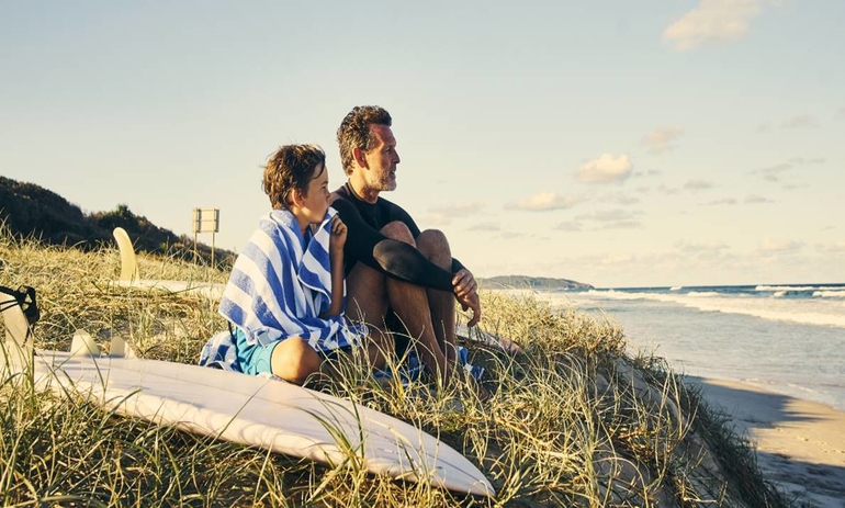 father and son sitting next to surfboard looking out to sea