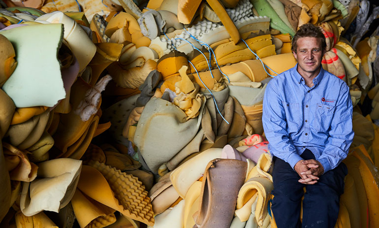 Man in a blue shift sitting on top a pile of foam mattresses