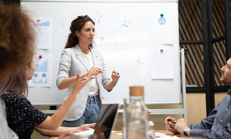 white woman with long brown hair wearing jeans and blazer standing in front of whiteboard giving presentation to group of people sat at table.