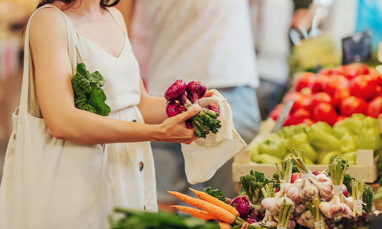 close up of lady in white dress shopping at a market, putting onions into a mesh bag she has brought