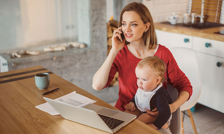 Woman sitting at the table in her kitchen with her laptop, speaking on her mobile while holding a baby on her knee