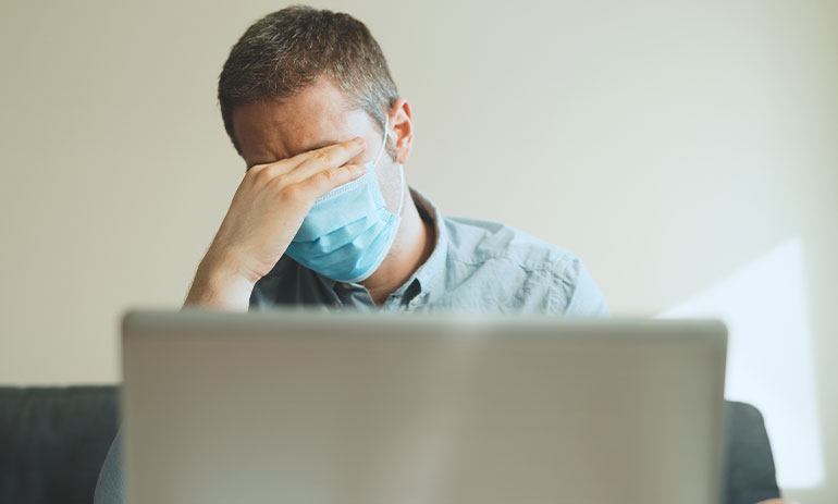 man looking tired wearing face mask sitting at laptop