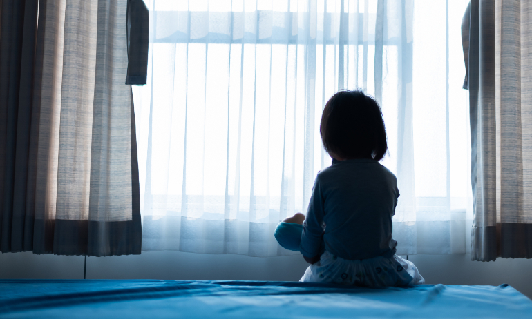 Little child sitting on bed playing with the doll in the dark room with light through curtain