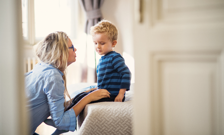 A young mother talking to her toddler son inside in a bedroom.