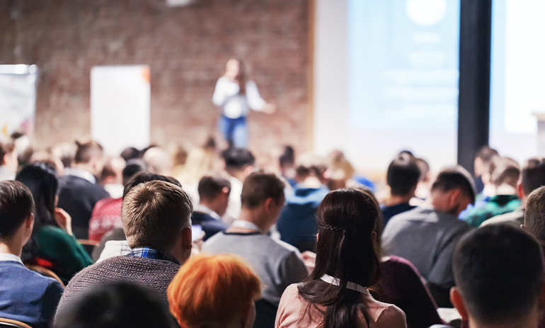 A group of people sit in an room listening to a woman speak