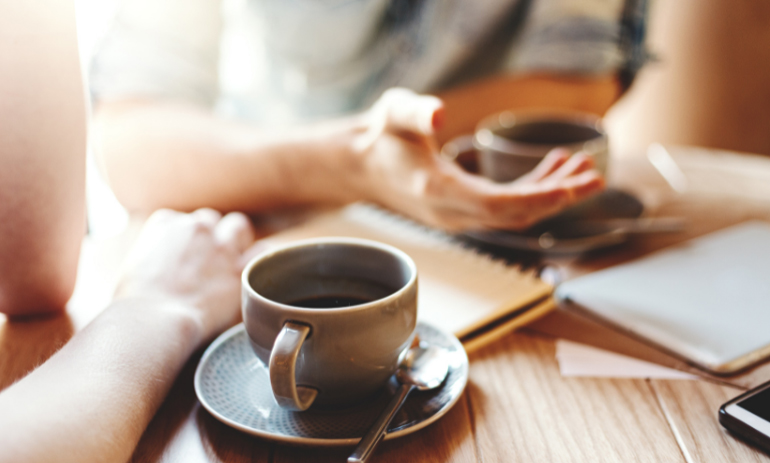 close up of table with two coffee cups and two people sitting having a chat