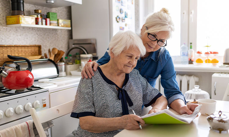 elderly woman sitting at a table looking at a file, another woman rests a hand on her shoulder