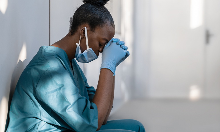 care worker in blue scrubs and a facemask sitting on the floor looking exhausted