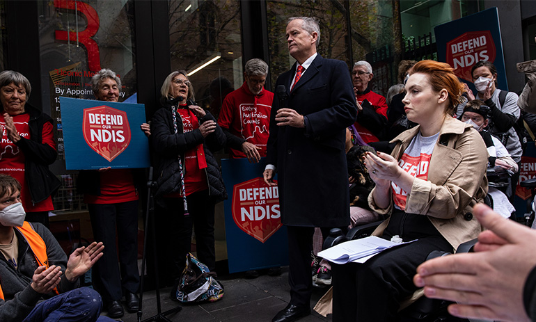 Bill Shorten, wearing a black suit and coat with a red tie, stands in the centre of a circle of people holding up blue and red signs that say 'defend our NDIS'. Some of the people are in wheelchairs.