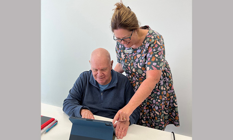 An elderly man wearing a blue shirt sits in front of an iPad at a table. A younger woman with brown hair, wearing a colourful dress, stands behind him and is pointing at the iPad. They are both smiling.