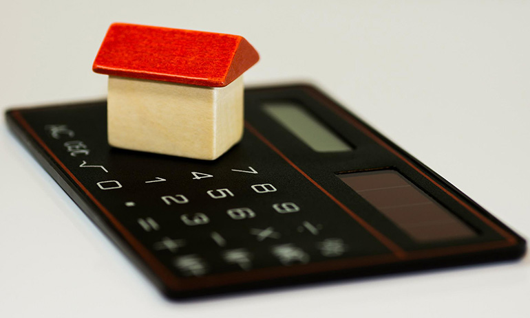 A black calculator with a model house sitting on top of it. The house has a red roof and beige walls.