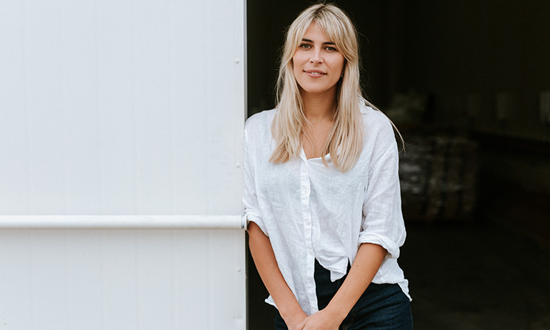 Julia Kay is looking at the camera with a small smile. She has long blonde hair and is wearing a white shirt and dark pants.